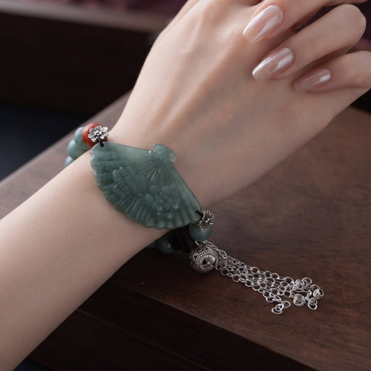 A close-up of a person's hand wearing a green jade-like bracelet with a silver chain tassel and a silver ring, resting on a dark wooden surface.