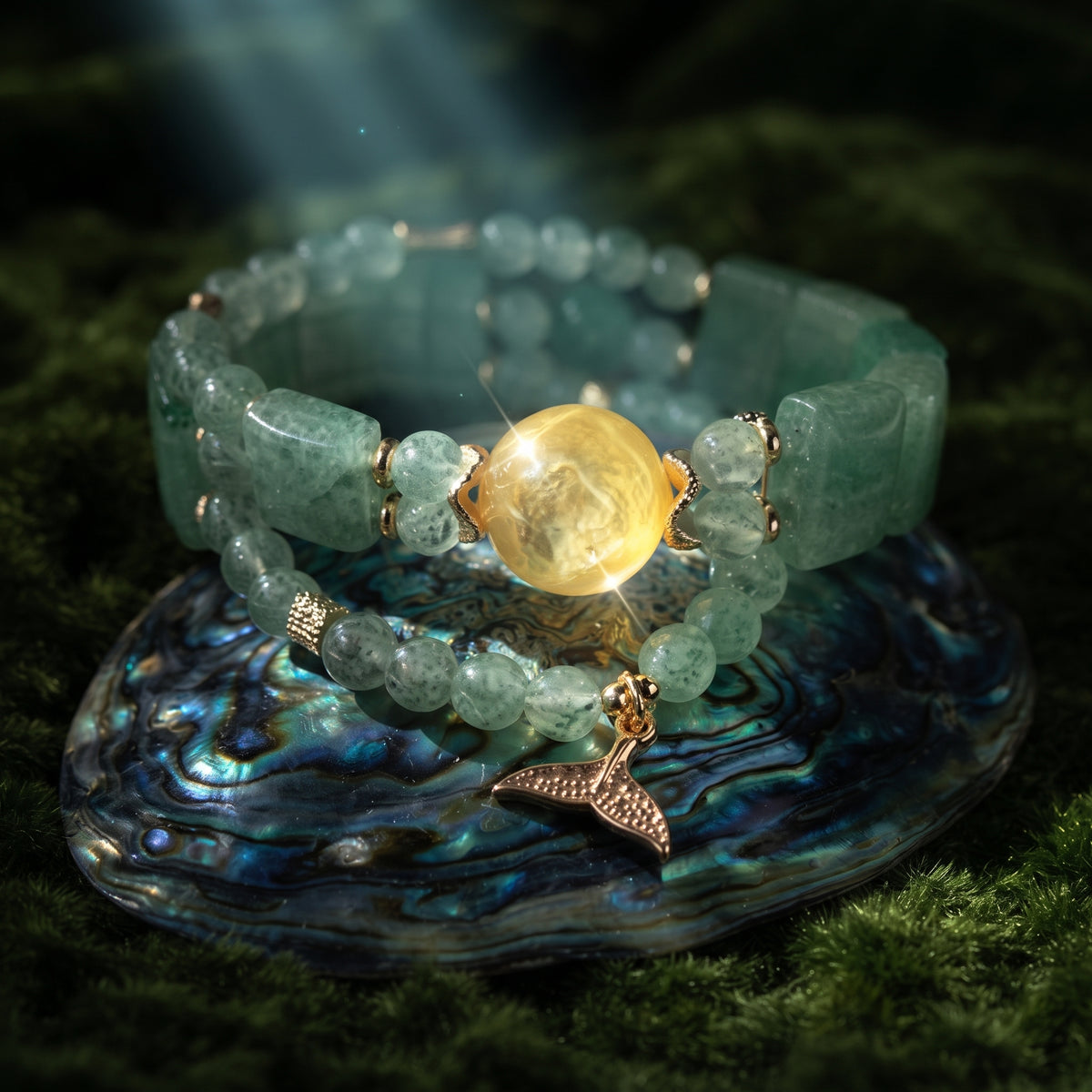 A beaded stretch bracelet made of light green stone beads with a central round yellow-gold bead and a dangling gold-toned mermaid tail charm, displayed on an abalone shell over a dark green textured surface.