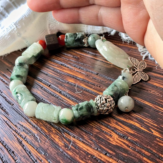 A beaded stretch bracelet made of green and white jade-like beads, carved cylindrical beads, a light green stone chip, a silver butterfly charm, and a silver elephant bead, held by a hand over a dark wood background.
