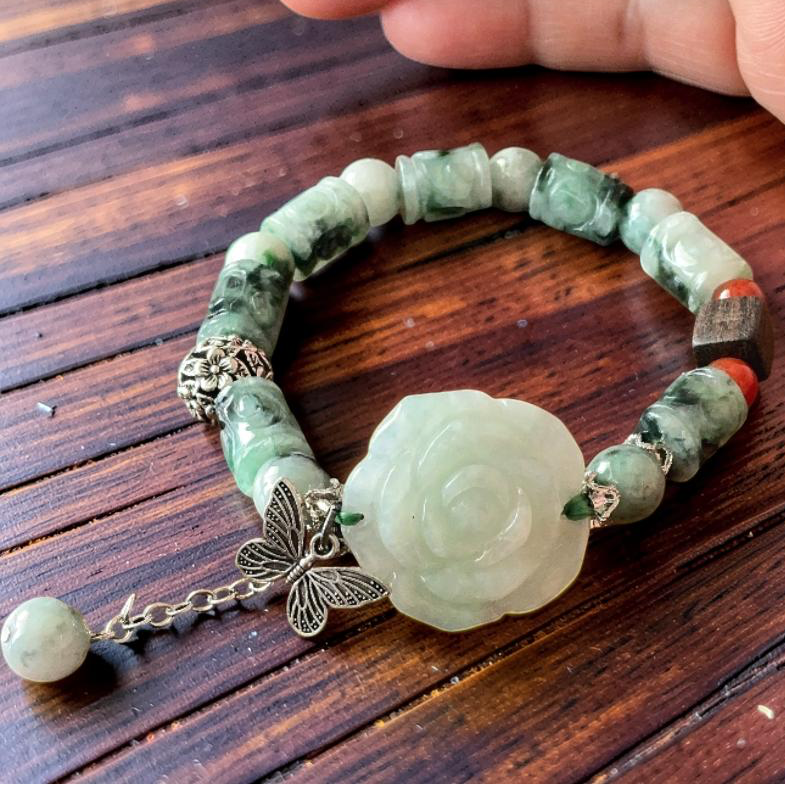 A beaded bracelet with green jade-like beads, a carved pale green rose centerpiece, and silver-tone charms including a butterfly, resting on a dark wooden surface with a hand held above it.
