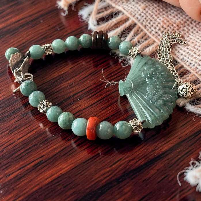 A beaded bracelet with light green jade-like beads, a carved green fan-shaped pendant, and small gold-toned charms, displayed on a dark wooden surface next to a person's hand.