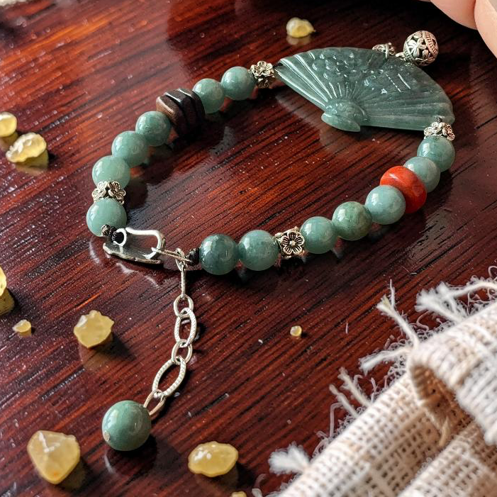 A beaded bracelet with light green jade-like beads, a carved green fan-shaped pendant, and small silver-toned spacer beads, displayed on a dark wooden surface with a hand nearby.