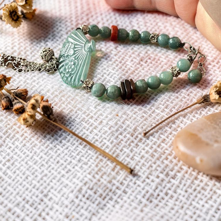 A handmade beaded bracelet with a carved light green jade pendant, green beads, and silver-toned metal accents, styled on a textured white linen cloth with dried flowers, a smooth stone, and a hand resting nearby.
