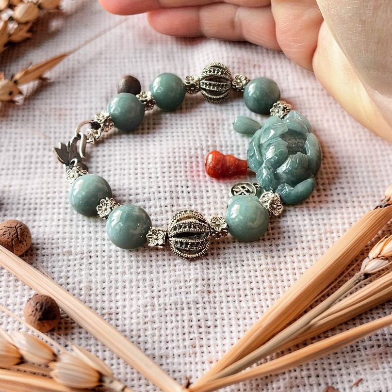 A beaded bracelet with turquoise-green round beads, silver filigree accents, and a carved green Buddha charm, styled on a textured cream fabric with dried wheat stalks and a hand partially visible.