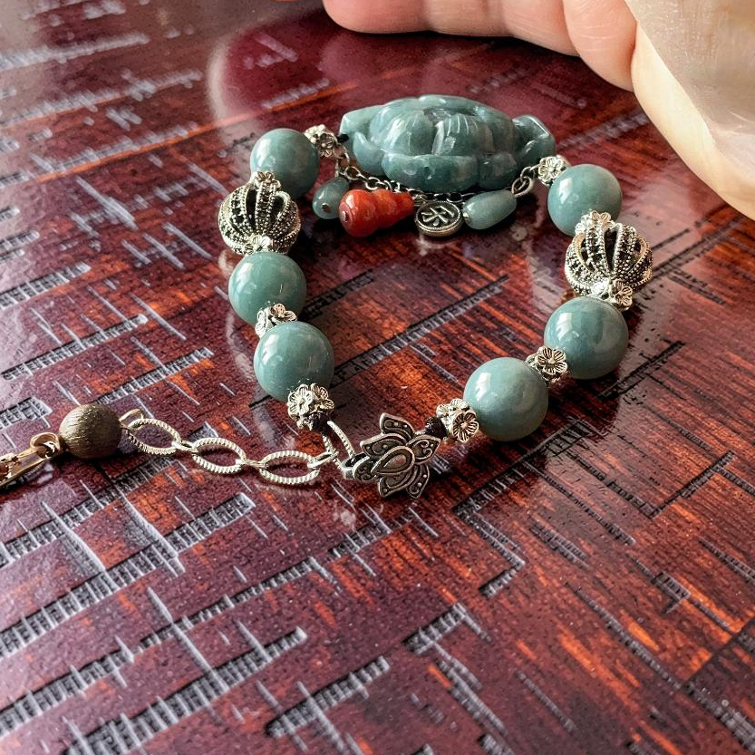 A beaded bracelet with light blue-green jade-like beads, a carved Buddha charm, and silver-toned metal accents, shown resting on a dark wooden surface with a hand partially visible at the top.
