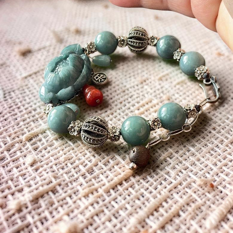 A handmade beaded bracelet with turquoise-green beads, a carved green jade lotus, and silver-toned filigree spacers, shown resting on a textured beige linen cloth with a hand holding the top right corner.