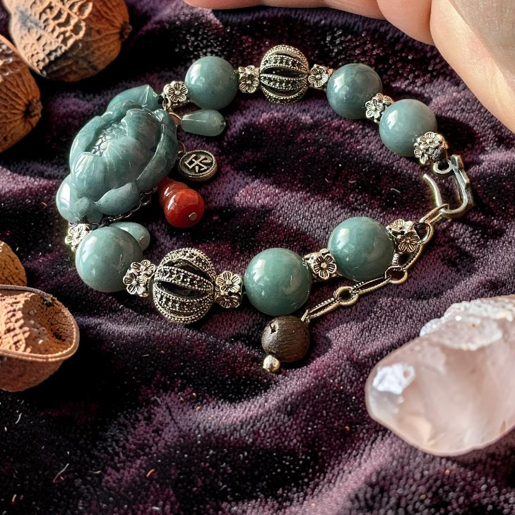 A beaded bracelet with turquoise-green beads, silver-toned filigree accents, and a carved green Buddha charm, styled on a dark purple velvet cloth with acorns, a crystal, and a hand nearby.