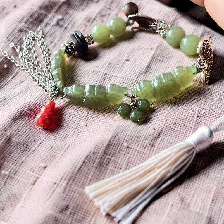A handmade beaded bracelet with light green jade-like beads, silver-toned metal details, a small red tassel charm, and a large white tassel, displayed on a textured beige fabric next to a hand.