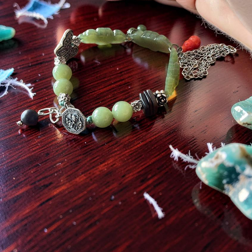 A handmade beaded bracelet with light green jade-like beads, a silver-toned elephant charm, and a small black bead, displayed on a dark wooden table next to a hand.