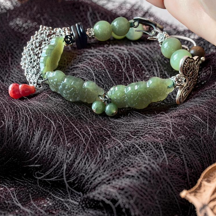 A handmade beaded bracelet with light green jade-like beads, silver-toned metal charms, and small red beads, displayed on a dark lace fabric with a hand holding the top edge.