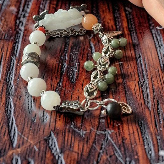 A close-up of a hand resting on a dark wooden surface next to a beaded bracelet. The bracelet features white and green beads, a carved white stone, and silver-toned metal charms.