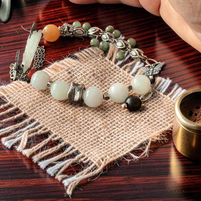A close-up of a hand resting on a dark wooden table beside a beaded bracelet and a small brass incense holder. The bracelet features white and green beads with silver-toned metalwork and a small orange accent bead, arranged on a piece of burlap fabric with dried greenery.