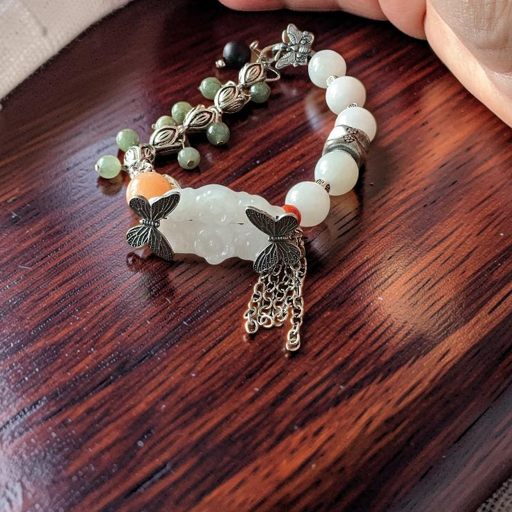 A beaded bracelet with a white jade-like central charm, butterfly charms, and green and white beads, resting on a dark wooden surface with a person's hands in the background.