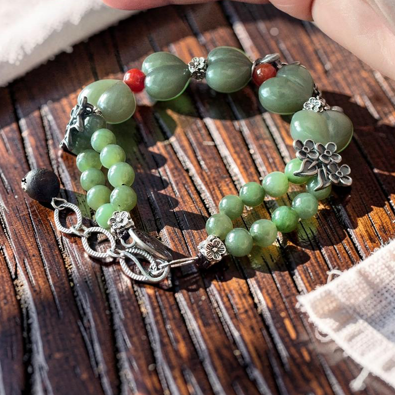 A beaded bracelet with light green jade-like beads, silver-toned metal charms, and small red and black accent beads, displayed on a dark wooden surface with a hand holding it for scale.