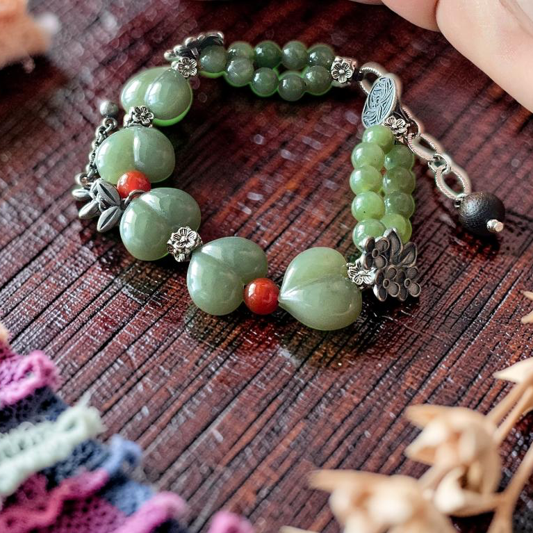 A handmade beaded bracelet made of green jade-like beads, small silver-toned metal charms, and a single red accent bead, displayed on a dark wooden surface with a hand resting nearby.