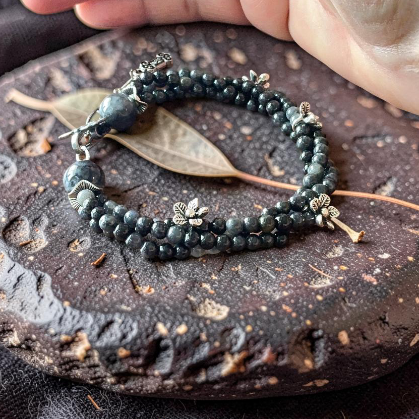 Dark jade double strand bracelet with silver floral charms and carved hulu pendant on stone surface