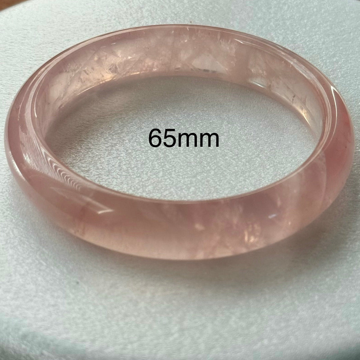 A translucent light pink stone bangle bracelet shown on a white textured display surface, with the text '65mm' overlaid near the center.