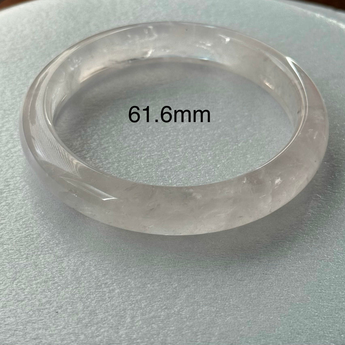 A translucent round bangle bracelet, likely made of quartz or crystal, displayed on a textured light gray surface with a black measurement overlay reading '61.6mm'.