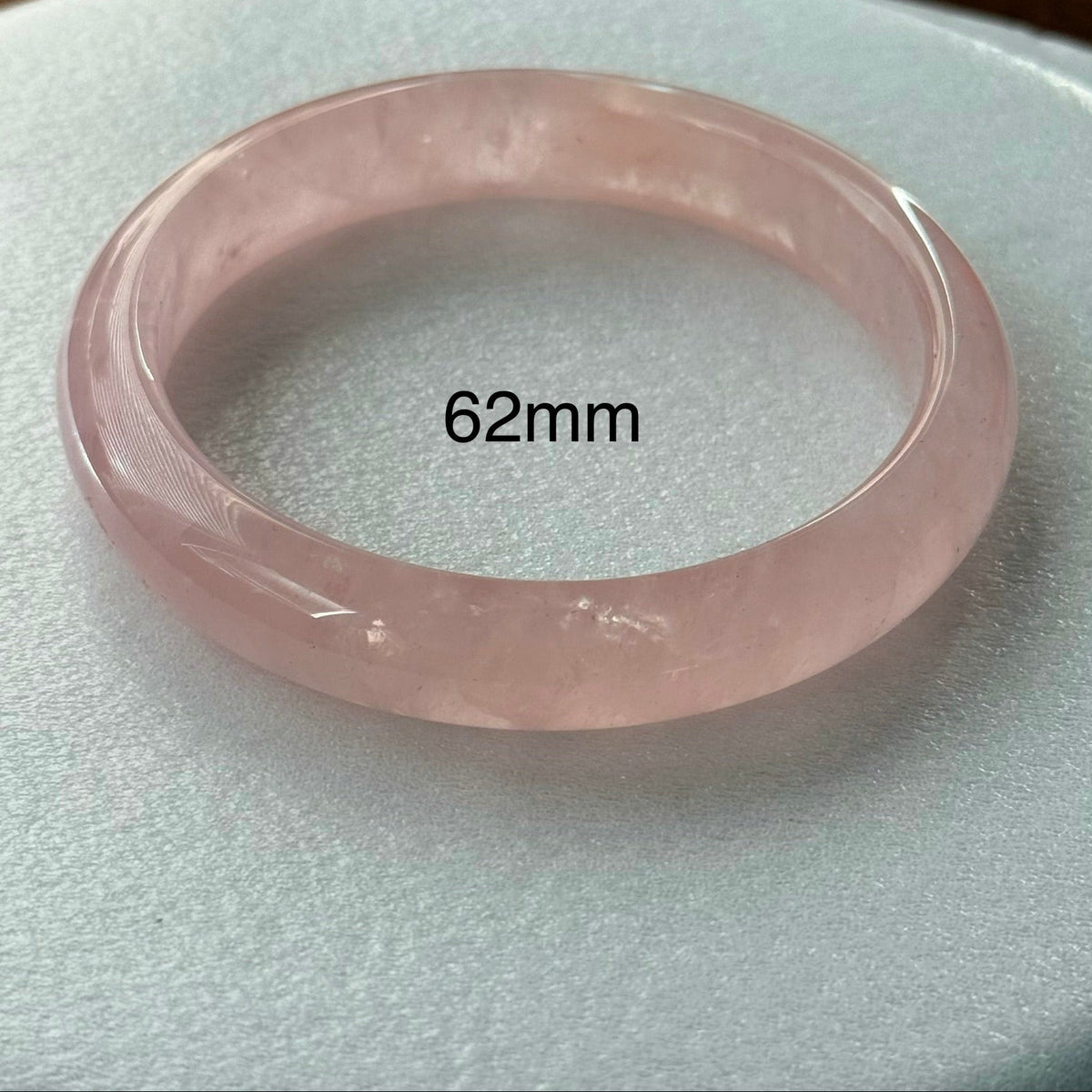 A translucent light pink jadeite bangle bracelet with a '62mm' size overlay, displayed on a textured white circular surface.