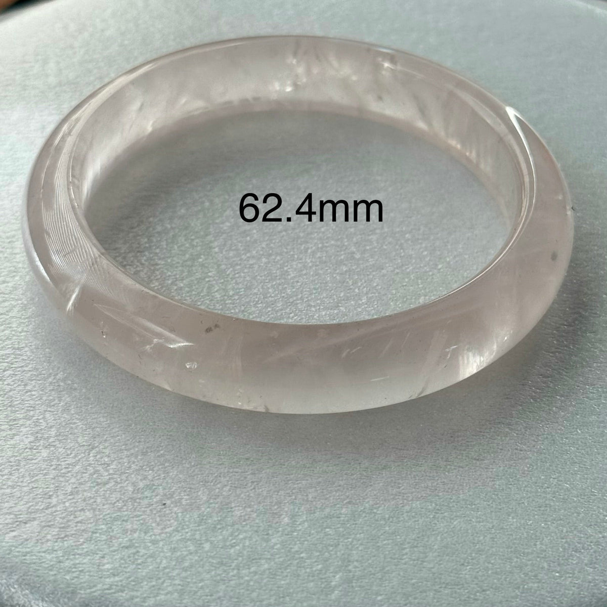 A translucent round bangle bracelet displayed on a light gray textured surface, with a black text overlay reading '62.4mm' indicating the diameter.