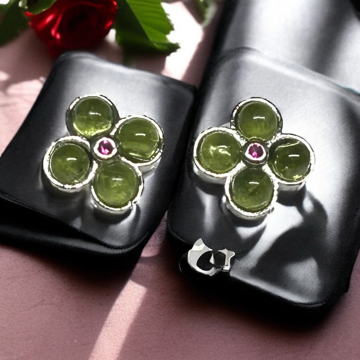 Green peridot flower-shaped earrings with a pink gemstone on black stands, placed on a pink surface.